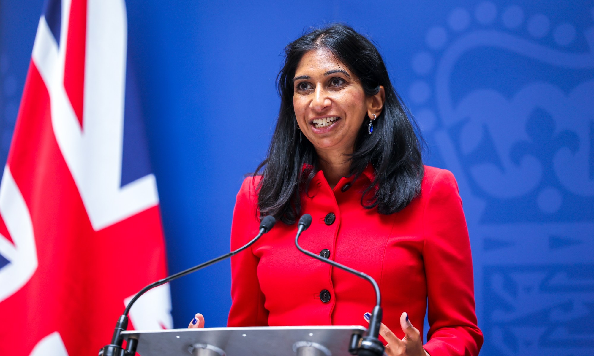 A person, Suella Bravemen, standing at a podium with microphones.