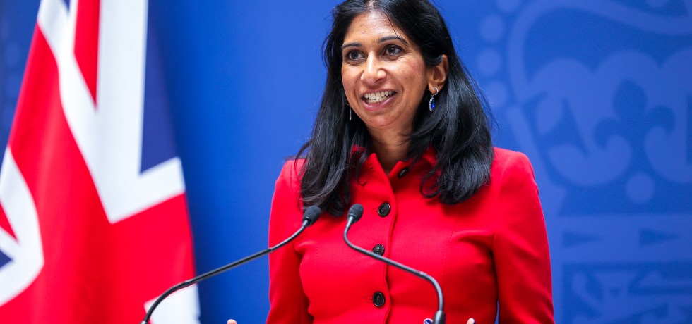 A person, Suella Bravemen, standing at a podium with microphones.