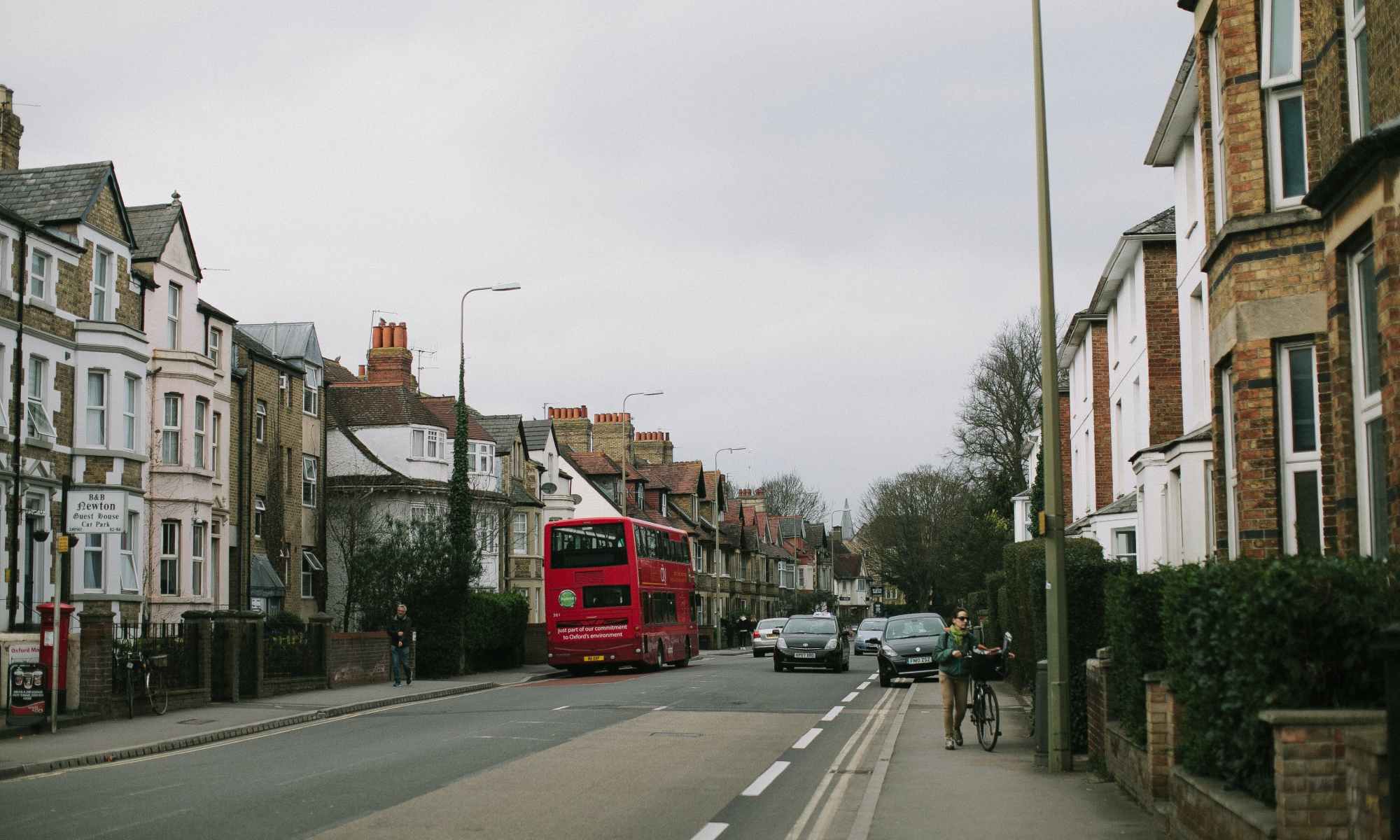 A red double decker bus on a street with cars and houses.